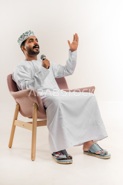 Working in presenting entertainment segments and artistic programs, expressions and gestures of happiness, portrait of a young Arab Gulf Omani man wearing a dishdasha and a kummah sitting on a comfortable chair holding a microphone in his hand, enjoying singing and presenting artistic talents, white background