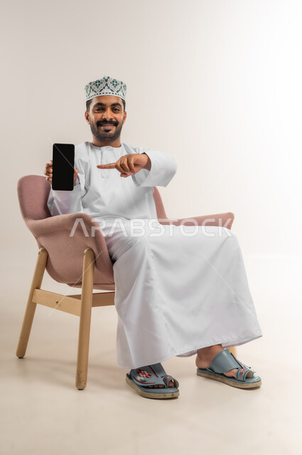 الاشارة الى عرض شاشة سوداء فارغة، دمج التكنولوجيا في الحياة اليومية، استخدام التقنيات والأجهزة الحديثة، بورتريه لرجل عربي خليجي عماني مبتسم يرتدي الدشداشة والكمة يجلس على كرسي مريح يقوم باستخدام الهاتف المحمول، خلفية بيضاء