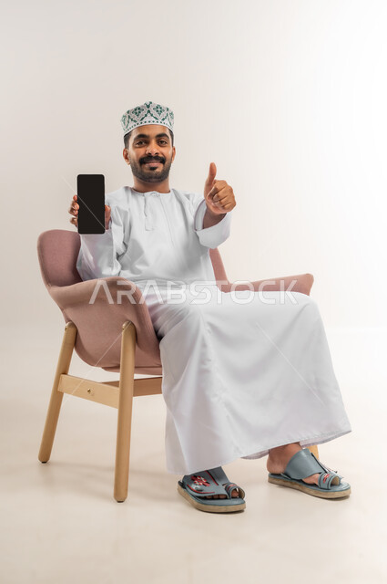 رفع اصبع الابهام بتعابير الموافقة والقبول والاعجاب، عرض شاشة سوداء فارغة، دمج التكنولوجيا في الحياة اليومية، استخدام التقنيات والأجهزة الحديثة، بورتريه لرجل عربي خليجي عماني مبتسم يرتدي الدشداشة والكمة يجلس على كرسي مريح يقوم باستخدام الهاتف المحمول، خلفية بيضاء