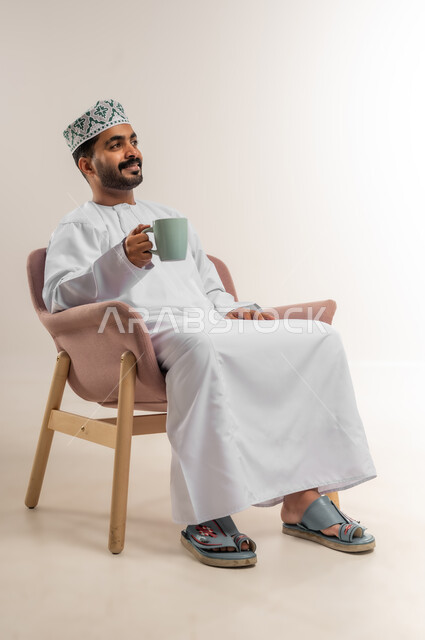 الاستمتاع بالمشروبات الساخنة، تناول الأعشاب الشتوية الدافئة، قضاء وقت الفراغ باحتساء المشروب المفضل, بورتريه لرجل عربي خليجي عماني مبتسم يرتدي الدشداشة والكمة يجلس على كرسي مريح يقوم بشرب القهوة بإيماءات السعادة والسرور، خلفية بيضاء