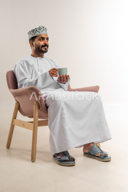 Enjoying hot drinks, eating warm winter herbs, spending free time sipping your favorite drink, portrait of a smiling Omani Gulf Arab man wearing a dishdasha and a turban sitting on a comfortable chair drinking coffee with gestures of happiness and pleasure, white background