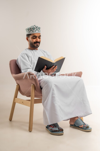 حب المطالعة والقراءة، تمضية أوقات الفراغ بالتعلم والتثقيف، بورتريه لرجل عربي خليجي عماني  يرتدي الدشداشه والكمة يجلس على اريكة مريحة يحمل بيده كتاب ينظر اليه بايماءات التركيز، الاهتمام بتصفح الكتب المفيدة، خلفية بيضاء