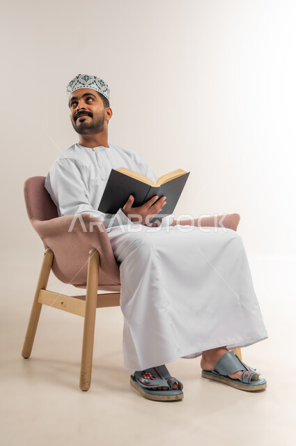حب المطالعة والقراءة، تمضية أوقات الفراغ بالتعلم والتثقيف، بورتريه لرجل عربي خليجي عماني  يرتدي الدشداشه والكمة يجلس على اريكة مريحة يحمل بيده كتاب ينظر الى مكان ما بايماءات التركيز، الاهتمام بتصفح الكتب المفيدة، خلفية بيضاء