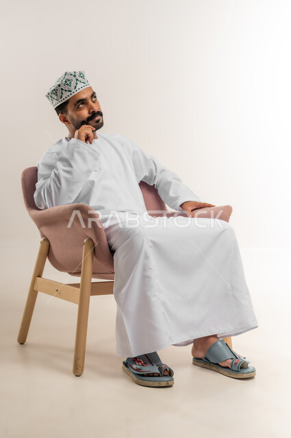 Finding a solution to a problem, contemplation and planning for the future, portrait of an Arab Gulf Omani man wearing a dishdasha and a kuma, putting his hand on his chin, looking somewhere with gestures of thinking and concentration, expressions of absent-mindedness and contemplation, white background