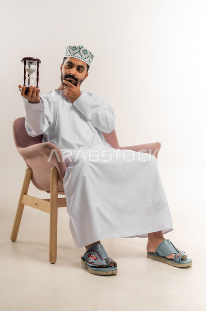 تعابير الانتظار والملل، مفهوم إدارة الأعمال وتنظيم الجدول الزمني, ادراك قيمة الوقت، بورتريه لرجل عربي خليجي عماني يرتدي الدشداشة والكمة يجلس على أريكة مريحة  يحمل ساعة رملية زجاجية (البنكام)، خلفية بيضاء