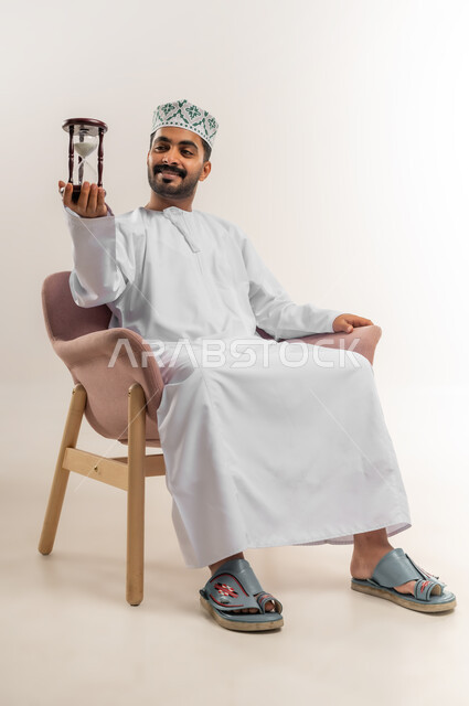 مفهوم إدارة الأعمال وتنظيم الجدول الزمني, ادراك قيمة الوقت، بورتريه لرجل عربي خليجي عماني يرتدي الدشداشة والكمة يجلس على أريكة مريحة  يحمل ساعة رملية زجاجية (البنكام)، خلفية بيضاء