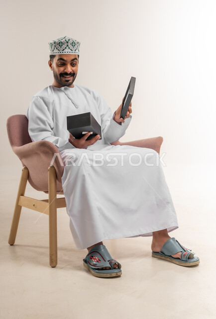 تقديم الهدايا بالاعياد والمناسبات الخاصة، مفهوم الشكر والامتنان، التعبير عن الحب والتقدير والاحترام، بورتريه لرجل عربي خليجي عماني يرتدي الدشداشة والكمة يجلس على أريكة مريحة يقوم بفتح صندوق الهدية بايماءات المفاجئة والصدمة، خلفية بيضاء