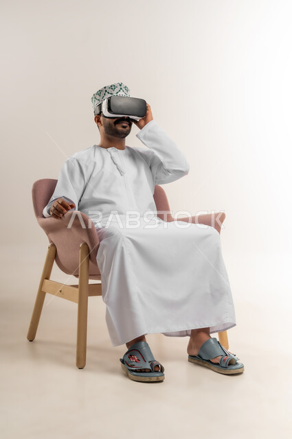المتعة والتسلية بالمشاهدة عبر نظارة الواقع الافتراضي، التطور التقني والتكنولوجي, بورتريه لشاب عربي خليجي عماني يرتدي الدشداشه والكمة يجلس على كرسي مريح ينظر من خلال النظارات ثلاثية الأبعاد, الاندماج ومحاكاة العالم الخيالي، متابعة الافلام الواقعيه، خلفيه بيضاء