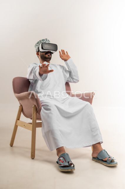 الاندماج ومحاكاة العالم الخيالي، المتعة والتسلية بالمشاهدة عبر نظارة الواقع الافتراضي، التطور التقني والتكنولوجي, بورتريه لشاب عربي خليجي عماني يرتدي الدشداشه والكمة يجلس على كرسي مريح ينظر من خلال النظارات ثلاثية الأبعاد, متابعة الافلام الواقعيه، خلفيه بيضاء