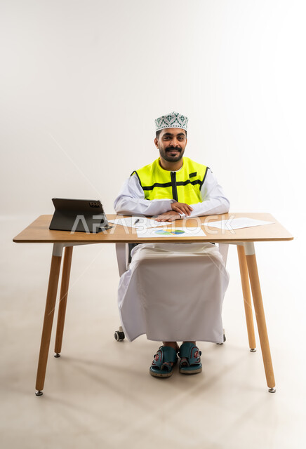 Studying graphs and statistics, following up on the construction plan through the technical device, working in the engineering sector, the concept of engineering and architectural construction, a portrait of an Arab Gulf Omani engineer wearing a dishdasha, a keffiyeh and a protective vest, holding a tablet in his hand, a profession and job for young people in the Sultanate of Oman, a white background