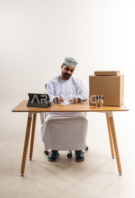 التسوق وطلب المنتجات عبر الإنترنت، توصيل الطلبات في الوقت المحدد وبشكل سليم، خدمة الشحن و التوصيل ذات الجودة العالية، بورتريه لشاب عربي خليجي عماني يرتدي الدشداشة والكمة يجلس على مكتبه امام صناديق كرتونية، تسجيل الطلبات من جهاز التابلت، خلفية بيضاء