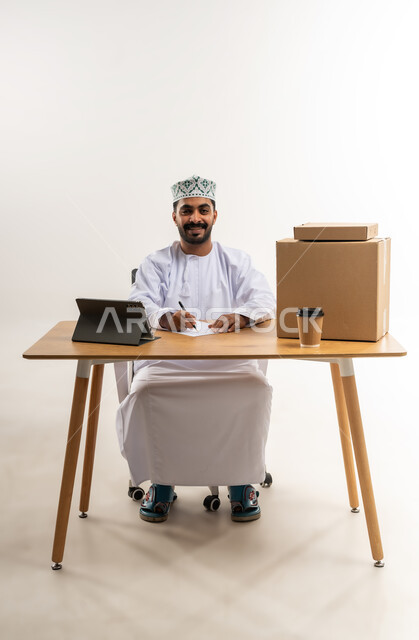 التسوق وطلب المنتجات عبر الإنترنت، توصيل الطلبات في الوقت المحدد وبشكل سليم، خدمة الشحن و التوصيل ذات الجودة العالية، بورتريه لشاب عربي خليجي عماني يرتدي الدشداشة والكمة يجلس على مكتبه امام صناديق كرتونية، تسجيل الطلبات من جهاز التابلت، خلفية بيضاء