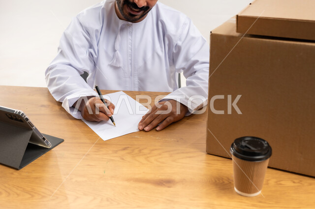 التسوق وطلب المنتجات عبر الإنترنت، توصيل الطلبات في الوقت المحدد وبشكل سليم، خدمة الشحن و التوصيل ذات الجودة العالية، بورتريه مقرب لشاب عربي خليجي عماني يرتدي الدشداشة والكمة يجلس على مكتبه امام صناديق كرتونية، تسجيل الطلبات من جهاز التابلت، خلفية بيضاء