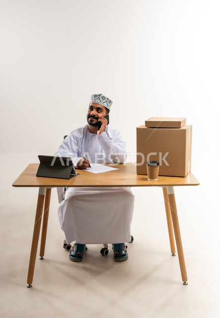 Responding to calls and inquiries, writing and recording customer requests, portrait of an Arab Gulf Omani man wearing a kummah and a dishdasha working in customer service at a freight and delivery company, documenting orders from a tablet, confirming reservations, managing the express delivery services process, white background