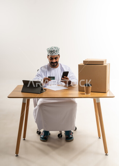 عمليات الدفع عن بعد, شراء المنتجات أونلاين عبر التابلت, مفهوم التسوق الالكتروني, بورتريه لشاب عربي خليجي عماني يرتدي الدشداشة و الكمة يجلس على الطاولة يمسك بيده الهاتف المحمول و بطاقة الائتمان البنكية, مفهوم خدمات التوصيل السريع, خلفية بيضاء
