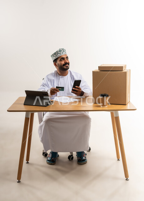 Contactless payments, online purchase of products via tablet, e-shopping concept, portrait of a young Omani Gulf Arab man wearing a dishdasha and a kummah sitting at a table holding a mobile phone and a bank credit card, express delivery services concept, white background