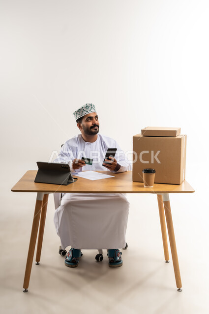 عمليات الدفع عن بعد, شراء المنتجات أونلاين عبر التابلت, مفهوم التسوق الالكتروني, بورتريه لشاب عربي خليجي عماني يرتدي الدشداشة و الكمة يجلس على الطاولة يمسك بيده الهاتف المحمول و بطاقة الائتمان البنكية, مفهوم خدمات التوصيل السريع, خلفية بيضاء