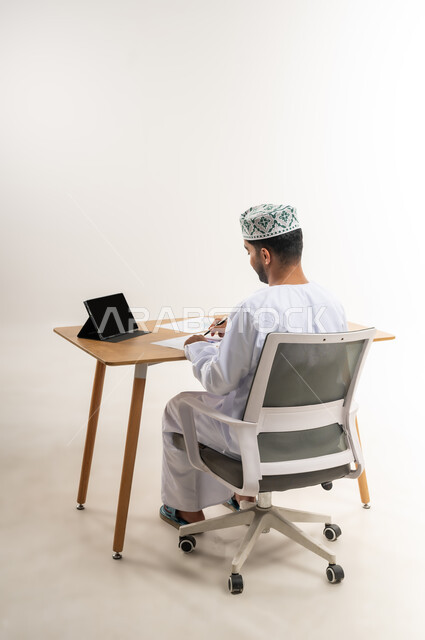 Business management using tablet, office profession and job in Omani companies, benefiting from modern technologies in the office and administrative sector, portrait from the back of an Arab Gulf Omani man wearing a dishdasha and a keffiyeh sitting behind an office table working hard and focused on the tablet, white background