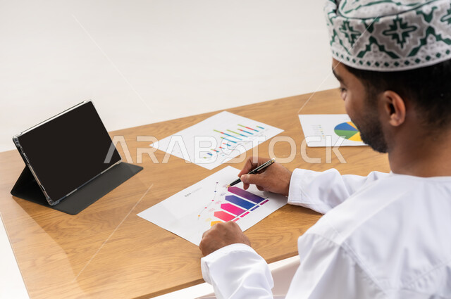 متابعة مخطط الرسم البياني للنمو الاقتصادي أونلاين، بورتريه مقرب من الخلف لرجل عربي خليجي عماني يرتدي الدشداشة والكمة يستخدم جهاز التابلت, العمل على أوراق الرسوم البيانية للسوق المالي، مراقبة الاقتصاد والمعلومات المالية، عرض شاشة سوداء فارغة، خلفية بيضاء