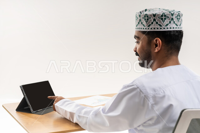 إدارة العمل باستخدام الجهاز اللوحي، مهنة ووظيفة مكتبية في الشركات العمانية، الاستفادة من التقنيات الحديثة في القطاع المكتبي والإداري، بورتريه مقرب من الخلف لرجل عربي خليجي عماني يرتدي الدشداشه والكمة يجلس خلف طاولة المكتب يعمل على التابلت بجد وتركيز، خلفية بيضاء