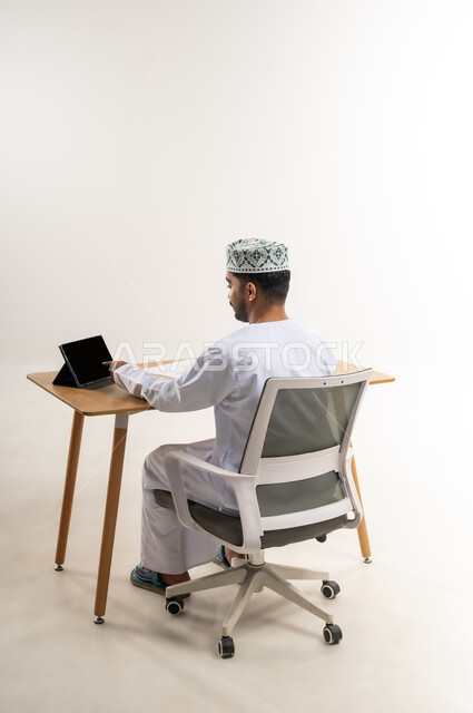 إدارة العمل عن بعد باستخدام الجهاز اللوحي، مهنة ووظيفة مكتبية في الشركات العمانية، الاستفادة من التقنيات الحديثة في القطاع المكتبي والإداري، بورتريه من الخلف لرجل عربي خليجي عماني يرتدي الدشداشه والكمة يجلس خلف طاولة المكتب يعمل على التابلت بجد وتركيز، خلفية بيضاء