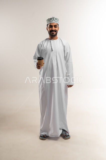 النظر الى الكاميرا بتعابير وايماءات السرور والسعاده, الاستمتاع بمذاق القهوه,  بورتريه لرجل عربي عماني خليجي يرتدي الدشداشه والكمه يحمل بيده كوب من المشروبات الساخنه, صورة الجسم بالطول الكامل, خلفيه بيضاء