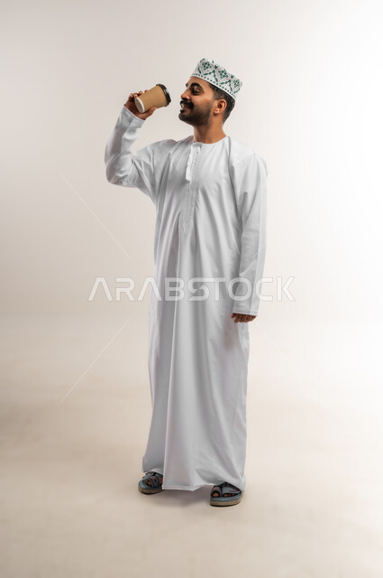 تعابير وايماءات السرور والسعاده, الاستمتاع بمذاق القهوه,  بورتريه لرجل عربي عماني خليجي يرتدي الدشداشه والكمه يحمل بيده كوب من المشروبات الساخنه, صورة الجسم بالطول الكامل, خلفيه بيضاء