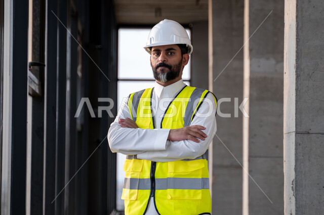 النظر الى الكاميرا بتعابير الثقة بالنفس والقوة، مهندس عربي خليجي سعودي يرتدي الثوب التقليدي والسترة وخوذة الحماية يقف بأيدي متقاطعة في موقع العمل، العمل في القطاع الهندسي في المملكة العربية السعودية، مفهوم ادارة المشاريع الهندسية والانشاءات، مهنة هندسية شبابية
