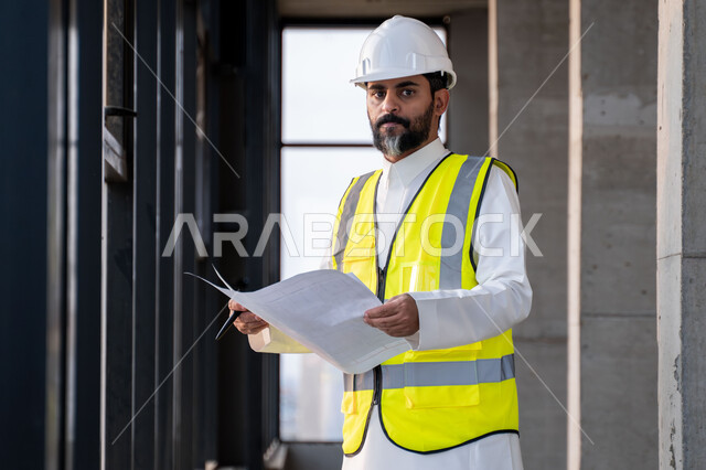  مفهوم ادارة المشاريع الهندسية والانشاءات، دراسة المخطط والمشروع على ارض الواقع، مهنة ووظيفة هندسية شبابية، مهندس عربي خليجي سعودي يرتدي الثوب التقليدي والسترة وخوذة الحماية يعمل في القطاع الهندسي في المملكة العربية السعودية