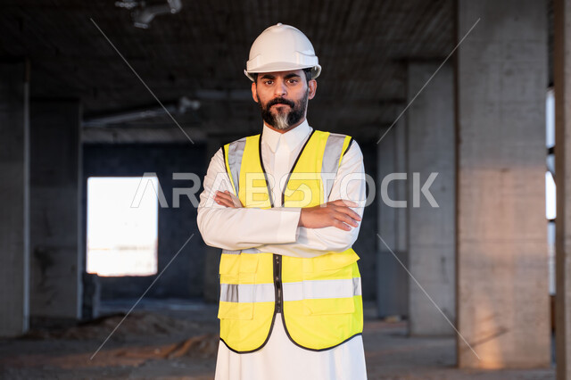 النظر الى الكاميرا بتعابير الثقة بالنفس والقوة، مهندس عربي خليجي سعودي يرتدي الثوب التقليدي والسترة وخوذة الحماية يقف بأيدي متقاطعة في موقع العمل، العمل في القطاع الهندسي في المملكة العربية السعودية، مفهوم ادارة المشاريع الهندسية والانشاءات، مهنة هندسية شبابية