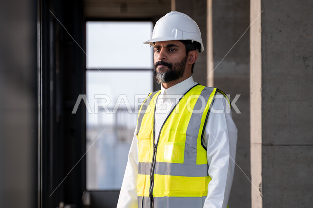 مهنة ووظيفة هندسية شبابية، مهندس عربي خليجي سعودي يرتدي الثوب التقليدي والسترة وخوذة الحماية يقف في موقع العمل، العمل في القطاع الهندسي في المملكة العربية السعودية، مفهوم ادارة المشاريع الهندسية والانشاءات