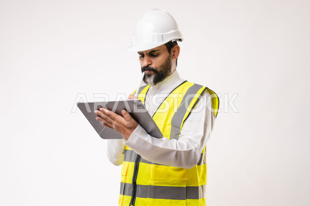 Using a modern and advanced technical device in the field of engineering, working in the Saudi engineering sector, the concept of engineering and construction project management, a youth profession and job, a portrait of a young Saudi Arabian Gulf engineer wearing traditional thobe, jacket and protective helmet working on a tablet, white background