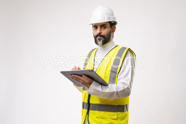 Using a modern and advanced technical device in the field of engineering, working in the Saudi engineering sector, the concept of engineering and construction project management, a youth profession and job, a portrait of a young Saudi Arabian Gulf engineer wearing traditional thobe, jacket and protective helmet working on a tablet, white background