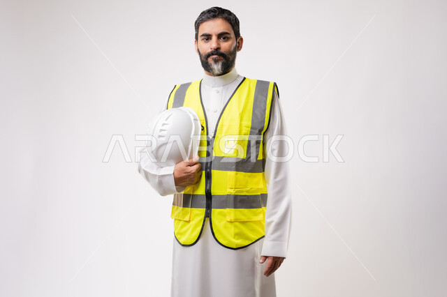 العمل في القطاع الهندسي في المملكة العربية السعودية، مفهوم ادارة المشاريع الهندسية والانشاءات، مهنة ووظيفة هندسية شبابية، بورتريه لمهندس عربي خليجي سعودي يرتدي الثوب التقليدي والسترة يمسك خوذة الحماية بيده يقف باستقامة بتعابير الحزم والجدية، خلفية بيضاء