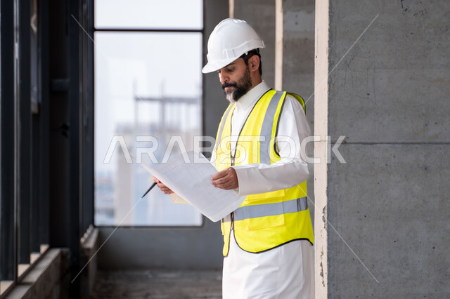 The concept of engineering and construction project management, studying the plan and the project on the ground, a youth engineering profession and job, a Saudi Arabian Gulf engineer wearing the traditional dress, jacket and protective helmet working in the engineering sector in the Kingdom of Saudi Arabia