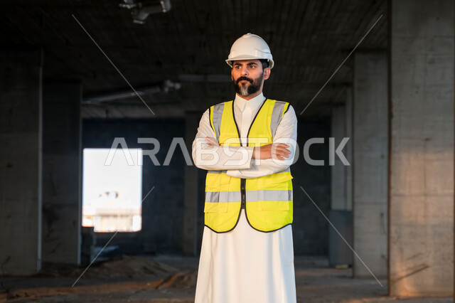 النظر الى الكاميرا بتعابير الثقة بالنفس والقوة، مهندس عربي خليجي سعودي يرتدي الثوب التقليدي والسترة وخوذة الحماية يقف بأيدي متقاطعة في موقع العمل، العمل في القطاع الهندسي في المملكة العربية السعودية، مفهوم ادارة المشاريع الهندسية والانشاءات، مهنة هندسية شبابية