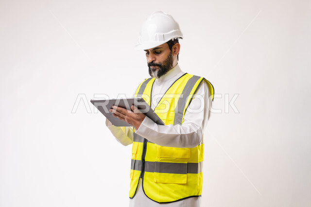 Using a modern and advanced technical device in the field of engineering, working in the Saudi engineering sector, the concept of engineering and construction project management, a youth profession and job, a portrait of a young Saudi Arabian Gulf engineer wearing traditional thobe, jacket and protective helmet working on a tablet, white background
