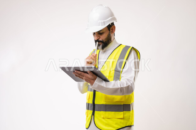 Expressions of thinking and concentration, using a modern and advanced technical device in the field of engineering, working in the engineering sector, the concept of engineering and construction project management, a youth profession and job, a portrait of a Saudi Arabian Gulf engineer wearing a traditional thobe, jacket and protective helmet working on a tablet, white background