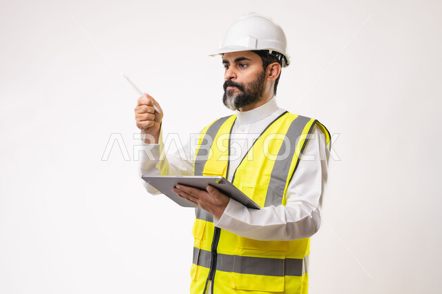 Using a modern and advanced technical device in the field of engineering, working in the Saudi engineering sector, the concept of engineering and construction project management, a youth profession and job, a portrait of a young Saudi Arabian Gulf engineer wearing traditional thobe, jacket and protective helmet working on a tablet, white background