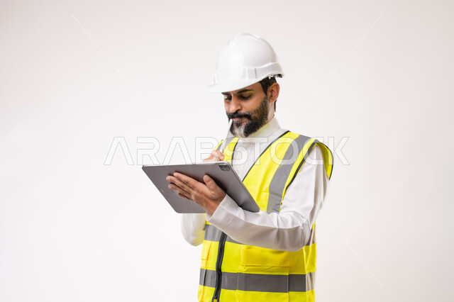 تعابير التفكير والتركيز، استخدام جهاز تقني حديث متطور في مجال الهندسة، العمل في القطاع الهندسي، مفهوم ادارة المشاريع الهندسية والانشاءات، مهنة ووظيفة شبابية، بورتريه لمهندس عربي خليجي سعودي يرتدي الثوب التقليدي والسترة وخوذة الحماية يعمل على جهاز التابلت، خلفية بيضاء