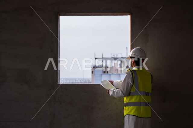 The concept of engineering and construction project management, studying the plan and the project on the ground, a youth engineering profession and job, a Saudi Arabian Gulf engineer wearing the traditional dress, jacket and protective helmet working in the engineering sector in the Kingdom of Saudi Arabia
