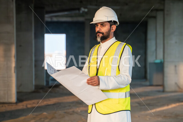 دراسة المخطط والمشروع  على ارض الواقع، مهنة ووظيفة هندسية شبابية، مهندس عربي خليجي سعودي يرتدي الثوب التقليدي والسترة وخوذة الحماية يعمل في القطاع الهندسي في المملكة العربية السعودية، مفهوم ادارة المشاريع الهندسية والانشاءات
