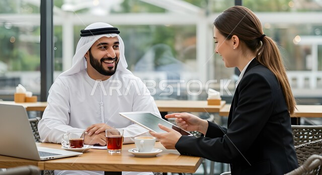 Cooperation in developing an appropriate business strategy, exchanging experiences and working methods, an external business meeting in one of the UAE cafes, a smiling Emirati Gulf Arab man wearing a ghutra and kandura sitting next to a foreign businesswoman, using a laptop to present projects and proposals