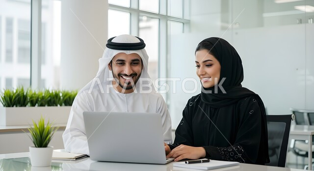 دمج التقنية والتكنولوجيا في الحياة العملية، رجل عربي خليجي اماراتي يرتدي الكندورة والغترة يجلس بجانب موظفة محجبة داخل المكتب في مقر الشركة، العمل على الحاسب المحمول، تبادل المعلومات والخبرات لتحقيق اهداف الشركة، بيئة العمل المريحة في الشركات الاماراتية