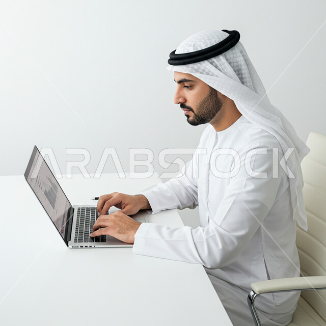 Working in offices and companies in the United Arab Emirates, performing tasks and office work inside the company headquarters, a side portrait of an Emirati Gulf Arab man wearing a kandura and ghutra sitting in front of his desk working on a laptop, integrating technology into practical life, white background
