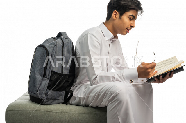 Portrait of a Saudi Arabian Gulf university student sitting next to his bookbag, enjoying reading the university book, studying for university exams, reviewing daily lessons, university education in the Kingdom of Saudi Arabia