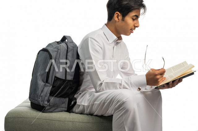 Portrait of a Saudi Arabian Gulf university student sitting next to his bookbag, enjoying reading the university book, studying for university exams, reviewing daily lessons, university education in the Kingdom of Saudi Arabia