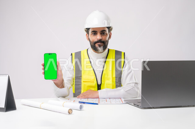 عرض شاشة خضراء فارغة على الهاتف المحمول، دمج التقنية والتكنولوجيا في الهندسة، العمل في القطاع الهندسي، ادارة المشاريع الهندسية والانشاءات، مهنة ووظيفة شبابية، بورتريه لمهندس عربي خليجي سعودي يرتدي خوذة الحماية والسترة يعمل على الحاسوب المحمول، كروما، خلفية بيضاء
