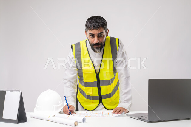 دمج التقنية والتكنولوجيا في مجال الهندسة، العمل في القطاع الهندسي في المملكة العربية السعودية، مفهوم ادارة المشاريع الهندسية والانشاءات، مهنة ووظيفة شبابية، بورتريه لمهندس عربي خليجي سعودي يرتدي الثوب التقليدي والسترة يعمل على الحاسوب المحمول، خلفية بيضاء