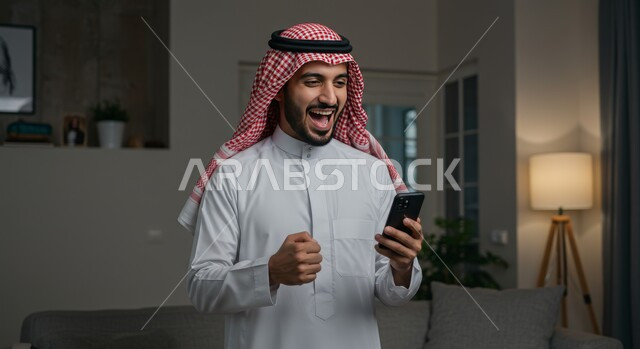 رؤية الاخبار المفرحة والسعيدة، تحقيق الاهداف والانجاز، تعابير النصر والفوز، صورة مقربة لرجل عربي خليجي سعودي يرتدي الشماغ والثوب التقليدي يمسك بيده الهاتف المحمول يرفع قبضة يده للاعلى بايماءات الحماس والسرور